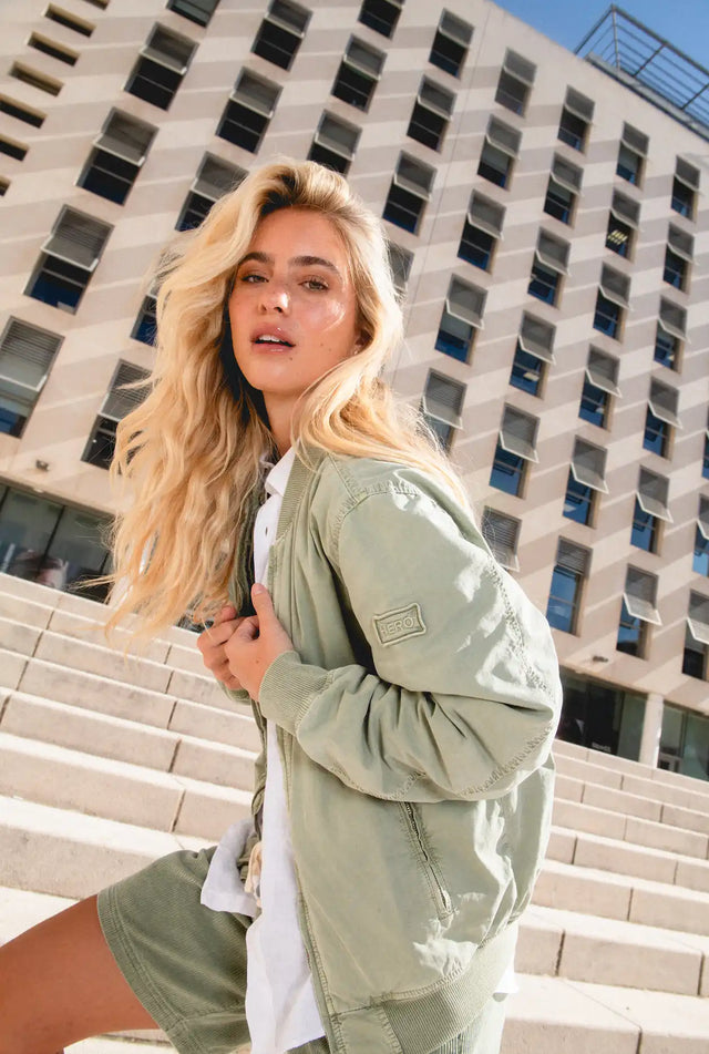 Une femme aux longs cheveux blonds portant une veste bomber vert clair, un t-shirt blanc et un short vert.