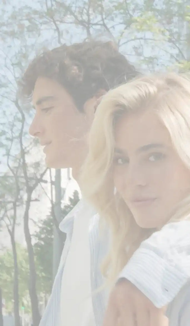 Un jeune couple, l'homme avec des cheveux bruns bouclés regardant vers la gauche, et la femme avec de longs cheveux blonds regardant droit devant, avec son bras autour de l'épaule de l'homme.