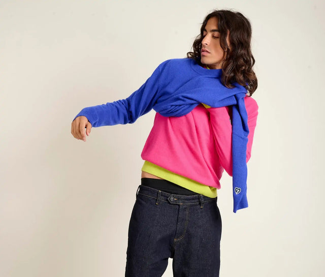 Une personne portant une chemise à manches longues rose vif, une sous-couche jaune néon, un jean bleu foncé décontracté, et une chemise à manches longues bleue drapée sur les épaules comme une écharpe.